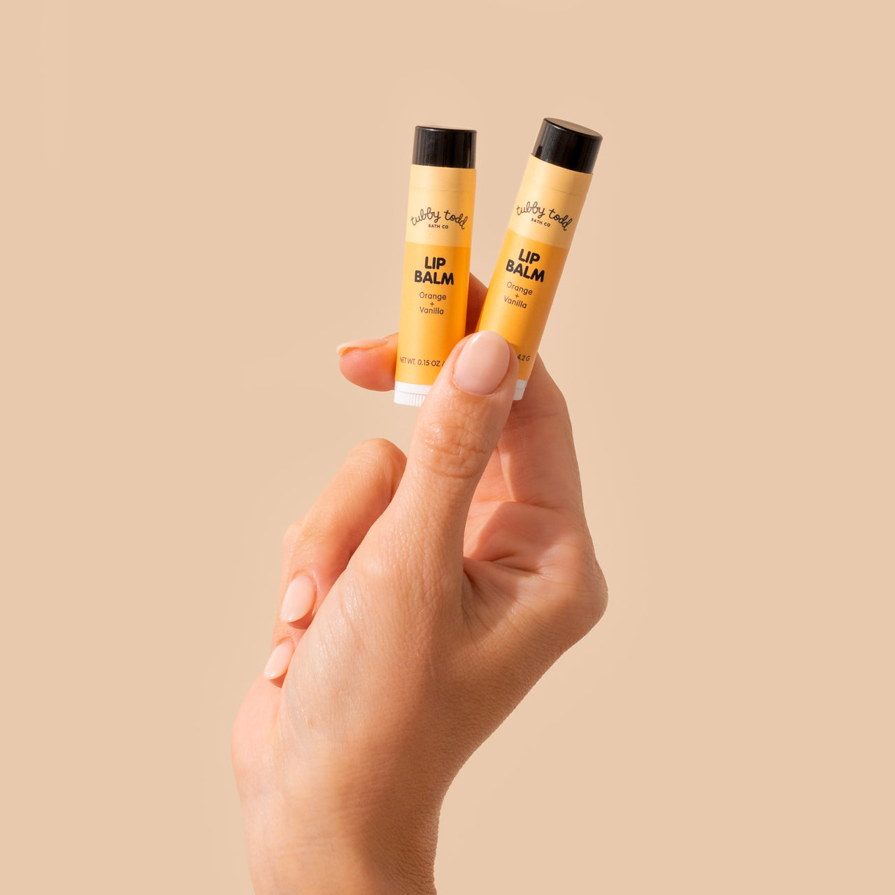 Hand holding two lip balm tubes against a beige background